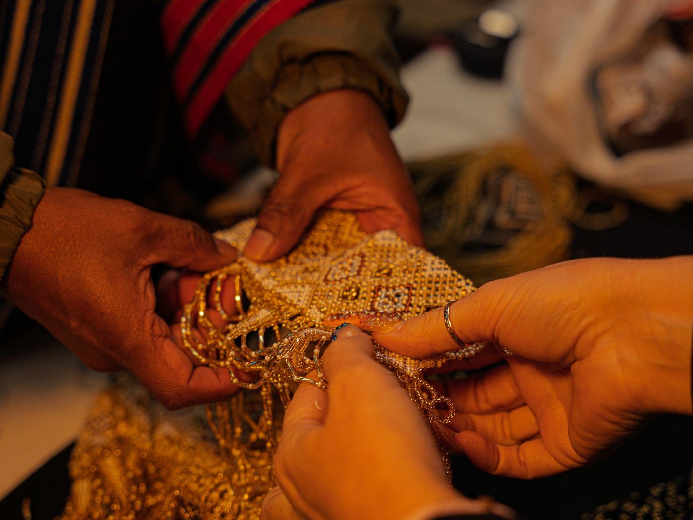 Two pairs of hands, an artisan's and Anika's, meeting over a gold-beaded piece in progress